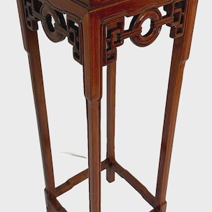 May include: A tall, square wooden pedestal stand with a polished reddish-brown finish. The top features a flat surface, while the upper section includes intricate geometric cutouts and circular motifs connecting the four slender, straight legs.