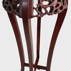 May include: A dark reddish-brown, carved wooden plant stand with a round top and four curved legs. The stand features intricate floral and bird carvings around the sides. The top appears to have a light-colored stone inlay.