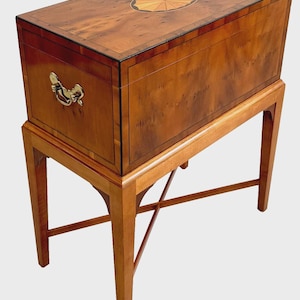 May include: A rectangular wooden chest with a polished burl wood finish, featuring a central oval inlay on the lid and brass side handles. The chest is supported by a matching wooden stand with four tapered legs and a cross-stretcher base.