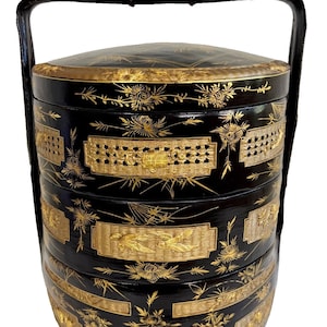 May include: A black and gold tiered food container with a woven bamboo design and floral accents. The container has a handle and is made of lacquered wood.