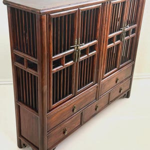 May include: A dark brown wooden cabinet featuring two sets of slatted doors with metal latch hardware. The lower section includes four drawers, each equipped with small metal pull handles. The piece has a rectangular frame and vertical slats.