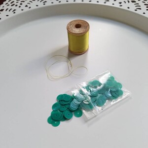 Peut inclure: Gros plan sur des fournitures de couture sur un plateau blanc. On distingue une bobine de fil jaune, du fil en vrac et un petit sachet de sequins turquoise. Le plateau est orné d'un bord ajouré.