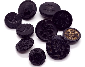 May include: A collection of black buttons with various embossed designs, including floral patterns, stars, and geometric shapes. The buttons vary in size and shape, with a few featuring gold accents. The buttons are arranged on a white background.