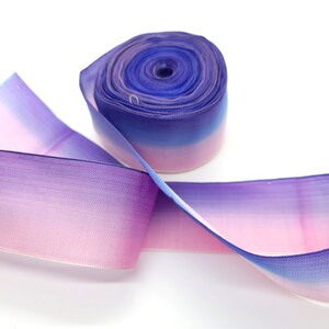 May include: A roll of ombre ribbon in shades of purple, pink, and blue. The ribbon is made of a sheer material, with a gradient effect from light pink to deep blue. The ribbon is unspooled, with a portion of it lying flat.
