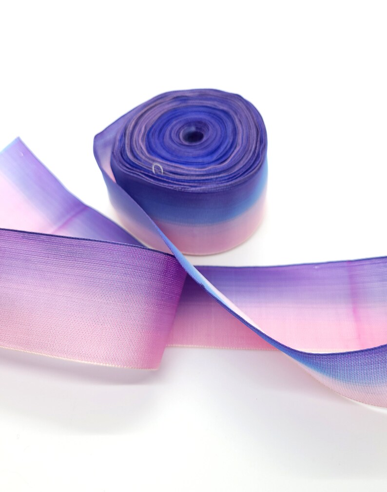 May include: A roll of ombre ribbon in shades of purple, pink, and blue. The ribbon is made of a sheer material, with a gradient effect from light pink to deep blue. The ribbon is unspooled, with a portion of it lying flat.