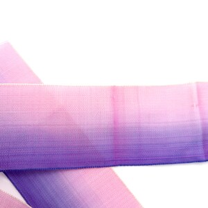 May include: A close-up shot of a length of ombre ribbon, fading from pink to purple. The ribbon has a woven texture and is set against a white backdrop. The colour gradient is smooth, creating a visually appealing effect. Suitable for crafting and gift wrapping.