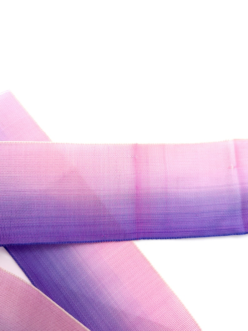 May include: A close-up shot of a length of ombre ribbon, fading from pink to purple. The ribbon has a woven texture and is set against a white backdrop. The colour gradient is smooth, creating a visually appealing effect. Suitable for crafting and gift wrapping.