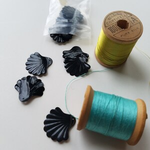 Pode incluir: Uma coleção de botões pretos em forma de concha, alguns em um saco plástico transparente, junto com dois carretéis de linha. Um carretel tem linha amarela, o outro tem linha turquesa. O rótulo do carretel amarelo diz "Belding Corticelli".