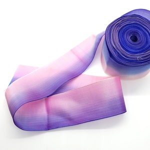 May include: A roll of ombre ribbon in shades of blue, purple, and pink. The ribbon is made of a sheer material and is unrolled on a white surface. The ribbon is likely used for crafts, gift wrapping, or other decorative purposes.