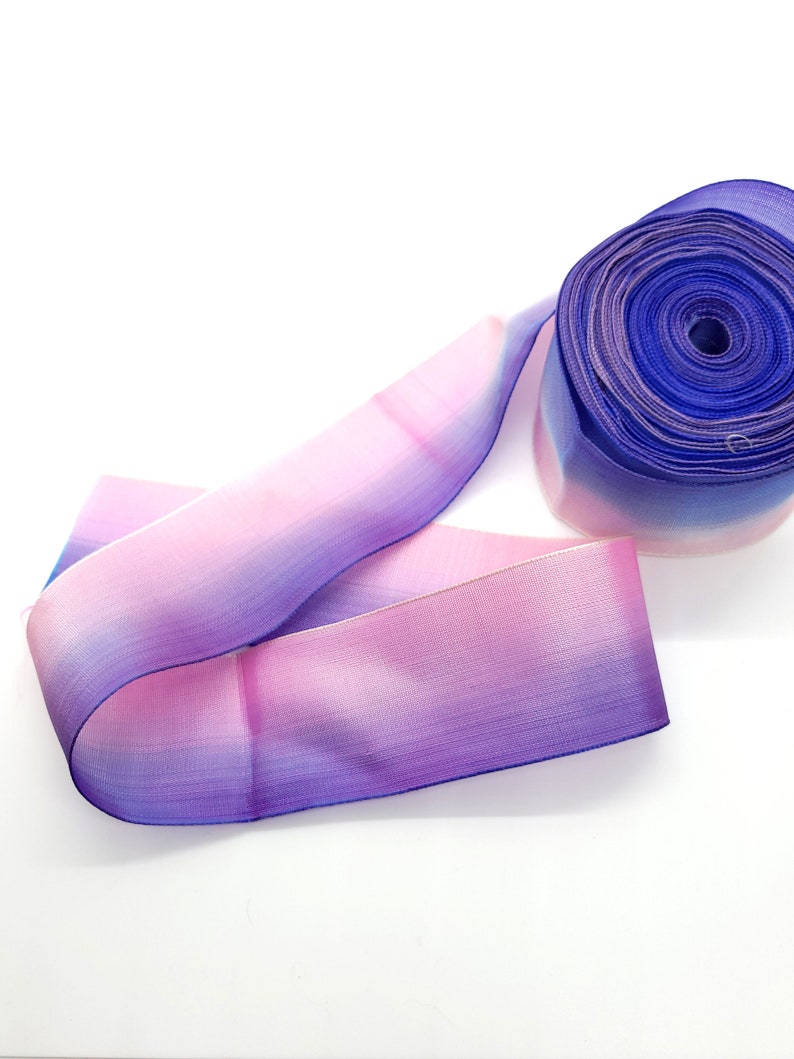 May include: A roll of ombre ribbon in shades of blue, purple, and pink. The ribbon is made of a sheer material and is unrolled on a white surface. The ribbon is likely used for crafts, gift wrapping, or other decorative purposes.