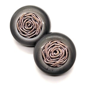 May include: Two dark gray, round stud earrings. Each earring features a rose design in the center, crafted from a lighter, metallic-looking material. The earrings are set against a white background.