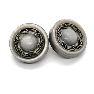 May include: Two gray buttons with a decorative silver-colored filigree design around a central gray dome. The buttons have a metallic sheen and are set against a white background.