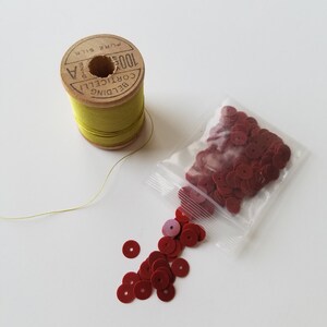 Könnte beinhalten: Ein Stillleben mit Nähzubehör. Eine Holzspule mit gelb-grünem Faden liegt neben einem kleinen Plastikbeutel mit roten Pailletten. Einige Pailletten sind aus dem Beutel gefallen. Die Spule ist mit "Belding Corticelli Pure Silk" beschriftet.