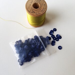 May include: A wooden spool of yellow thread with the text "PURE SILK" and "BELDING CORTICELLI" printed on it. A small plastic bag filled with navy blue sequins is next to the spool. A few sequins are scattered around the bag.