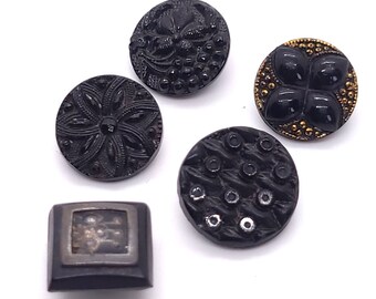 May include: A collection of five black buttons with various designs. The buttons include floral, geometric, and textured patterns. One button has a square shape with a silver-toned border and a detailed center.