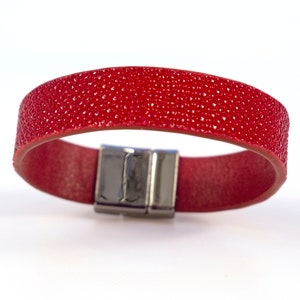 Puede incluir: Un brazalete de cuero rojo con un cierre magnético plateado. El brazalete tiene una superficie texturizada.