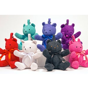 May include: Eight plush teddy bear keychains in various colors, including red, white, teal, purple, black, and pink. Each bear has a small bow tied around its neck.