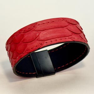 Puede incluir: Una pulsera de cuero roja con un patrón texturizado en forma de escamas. La pulsera tiene un interior negro y un cierre magnético negro. Las costuras rojas acentúan los bordes de la pulsera.
