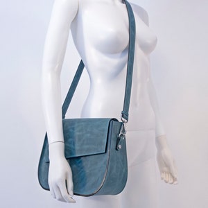May include: A blue leather shoulder bag with a flap closure and a silver buckle. The bag is worn on a white mannequin.