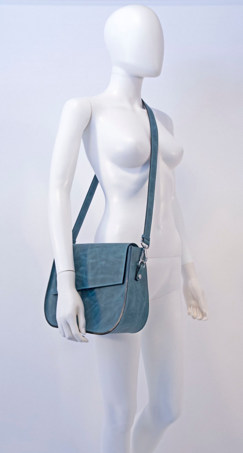 May include: A blue leather shoulder bag with a flap closure and a silver buckle. The bag is worn on a white mannequin.