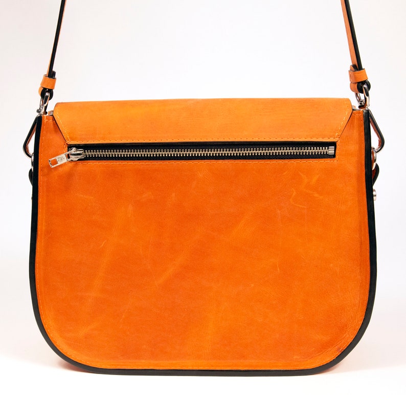 K&ouml;nnte beinhalten: Eine braune Ledertasche mit schwarzem Besatz und einem silbernen Rei&szlig;verschluss. Die Tasche hat einen langen Riemen und einen Klappverschluss.