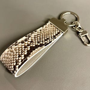 May include: A snakeskin-patterned keychain with a silver-tone metal ring and clasp. The keychain strap features a reptile print in shades of brown, tan, and white, with a white border and gray backing. The metal hardware adds a touch of elegance.