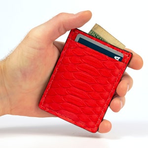 Puede incluir: Una cartera de piel roja de serpiente con una ranura para tarjetas y un bolsillo para billetes. La cartera est&aacute; siendo sostenida en una mano.