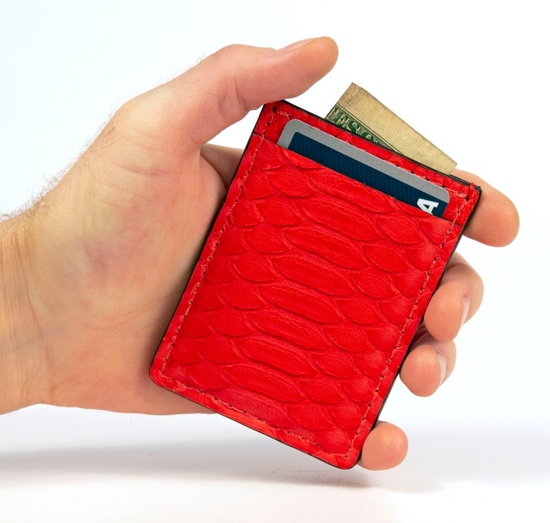 Puede incluir: Una cartera de piel roja de serpiente con una ranura para tarjetas y un bolsillo para billetes. La cartera est&aacute; siendo sostenida en una mano.
