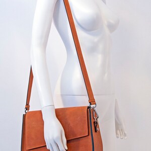 K&ouml;nnte beinhalten: Eine braune Lederschultertasche mit Klappenverschluss und einem silbernen Verschluss. Die Tasche hat einen langen verstellbaren Riemen.