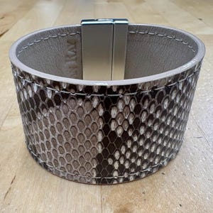 May include: A wide cuff bracelet with a snakeskin pattern in shades of brown, gray, and white. The bracelet has a light beige interior and a silver-colored magnetic clasp. The stitching is visible along the edges.