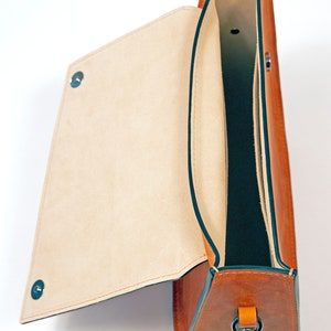 K&ouml;nnte beinhalten: Eine braune Ledertasche mit einem Klappdeckel und einem dunkelgr&uuml;nen Futter. Die Tasche hat einen einzigen Riemen mit einer Metallschnalle.