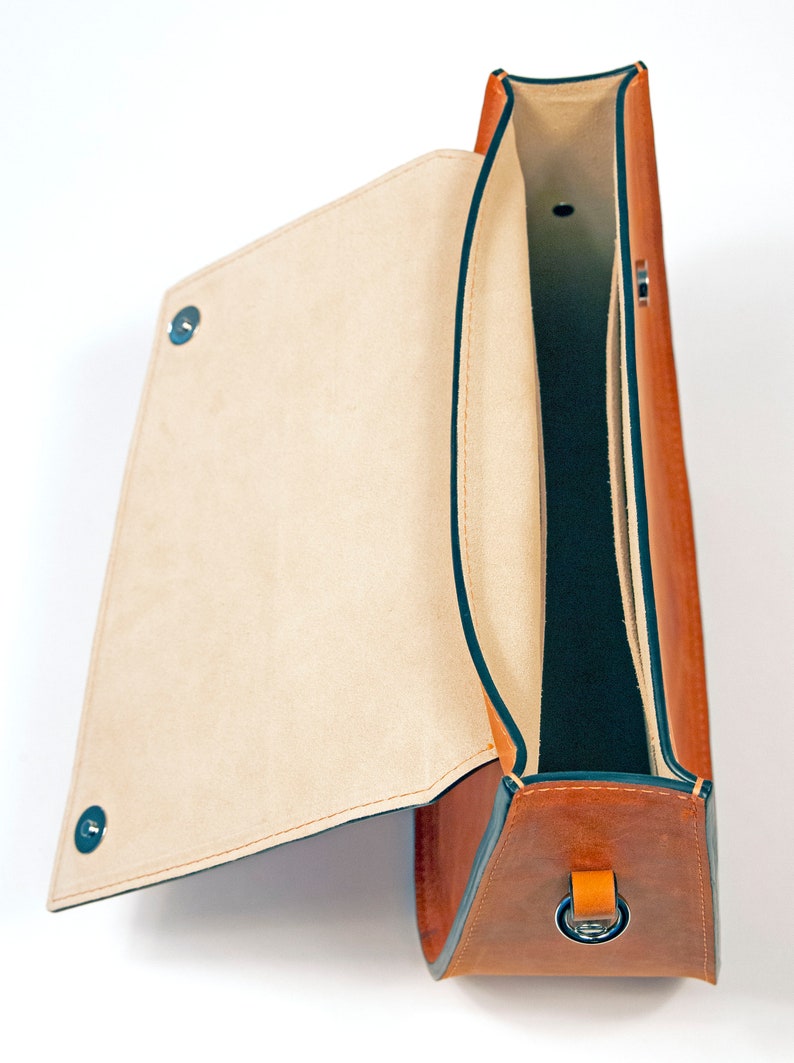 K&ouml;nnte beinhalten: Eine braune Ledertasche mit einem Klappdeckel und einem dunkelgr&uuml;nen Futter. Die Tasche hat einen einzigen Riemen mit einer Metallschnalle.
