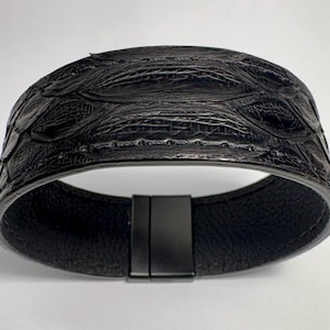 May include: A black leather cuff bracelet with a textured, scale-like pattern on the top surface. The bracelet has a magnetic clasp and a smooth interior. The cuff is approximately 2 inches wide.