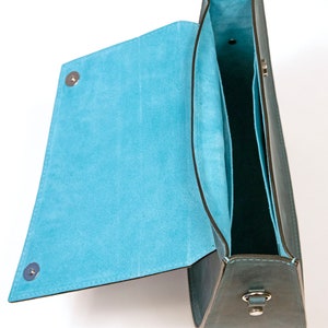 May include: A teal leather handbag with a flap top and a blue suede interior. The bag has a silver clasp and a silver strap.
