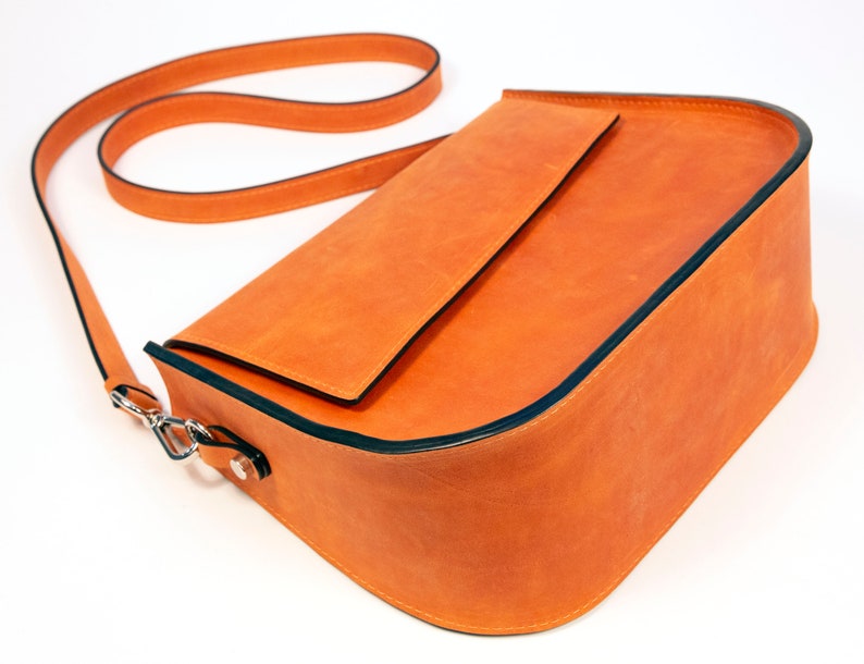 K&ouml;nnte beinhalten: Eine leuchtend orangefarbene Lederschultertasche mit schwarzem Besatz und einem langen Trageriemen. Die Tasche hat einen Klappenverschluss und ein kleines Fach auf der Vorderseite.