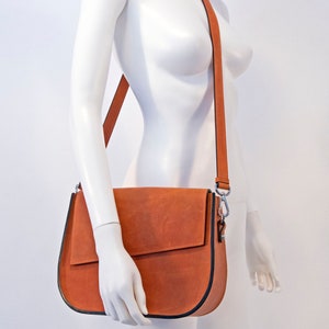 K&ouml;nnte beinhalten: Eine braune Lederschultertasche mit schwarzem Besatz und einem silbernen Verschluss. Die Tasche wird von einer wei&szlig;en Schaufensterpuppe getragen.