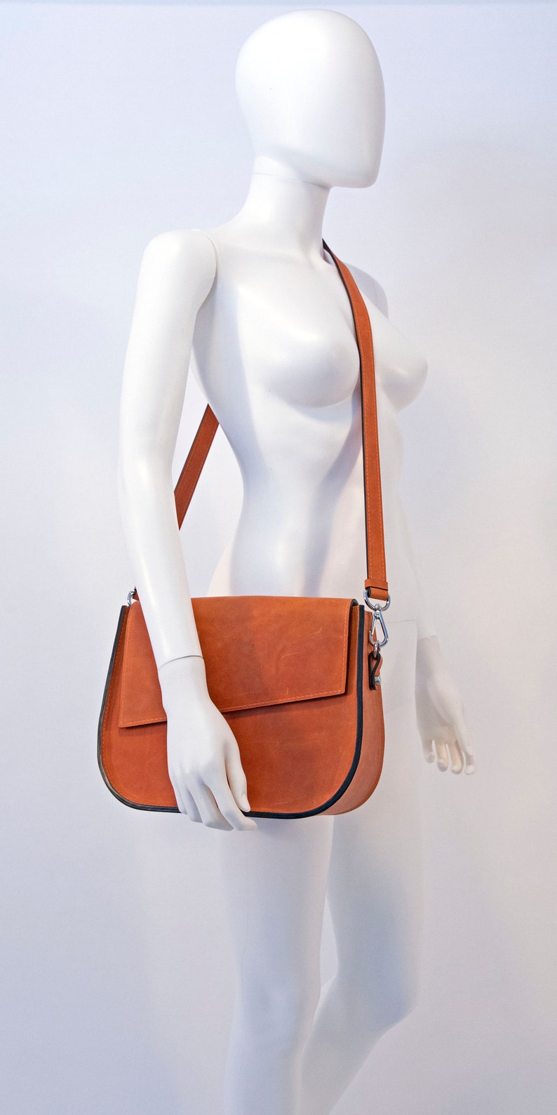 K&ouml;nnte beinhalten: Eine braune Lederschultertasche mit schwarzem Besatz und einem silbernen Verschluss. Die Tasche wird von einer wei&szlig;en Schaufensterpuppe getragen.