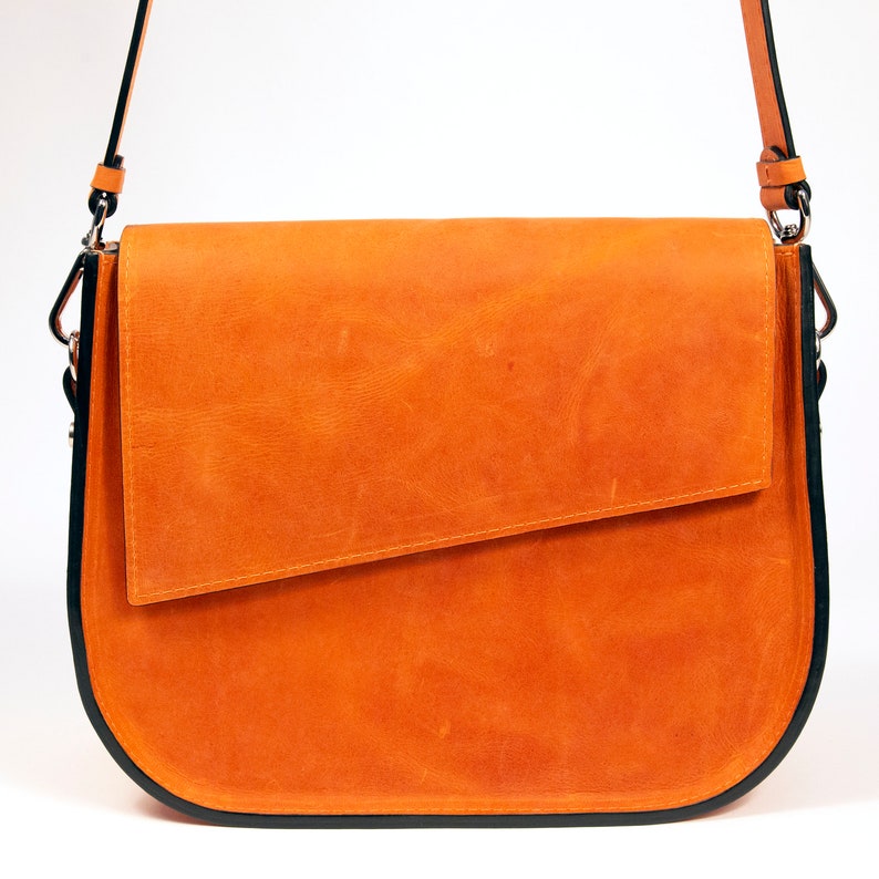 K&ouml;nnte beinhalten: Eine braune Ledertasche mit schwarzem Besatz und einem Klappverschluss. Die Tasche hat einen langen Riemen zum Tragen &uuml;ber der Schulter.