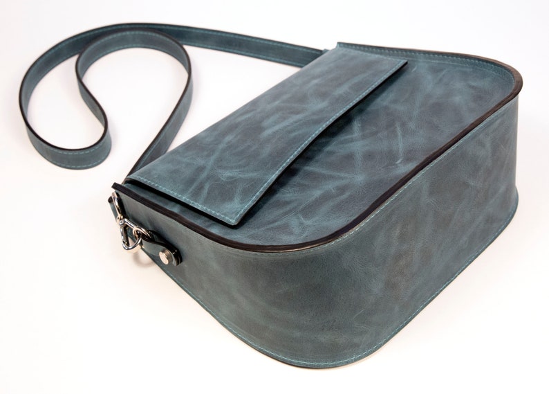 May include: A teal blue leather shoulder bag with a flap closure and a small pocket on the front. The bag has a long adjustable strap.