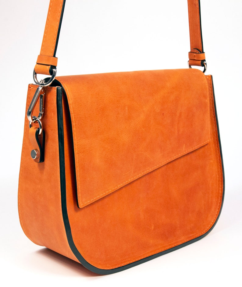 K&ouml;nnte beinhalten: Eine braune Leder-Umh&auml;ngetasche mit schwarzem Besatz. Die Tasche hat einen Klappenverschluss und einen langen, verstellbaren Riemen.