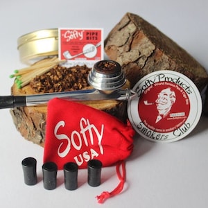 Puede incluir: Una pipa de metal plateado con una bolsa roja, cuatro boquillas de pipa negras, una lata de tabaco y una pegatina roja y blanca que dice "Softy Products - Original World Famous Pipe & Cigar Bits - Smokers Club".