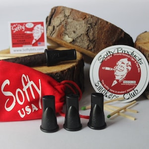 May include: Three black cigar mouthpieces in a red pouch with the word "Softy USA" printed on it. The mouthpieces are on a white surface with a wooden log and a red and white sticker that says "Softy Products Original World Famous Cigar & Pipe Co. Smokers Club" in the background.