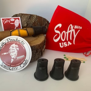 May include: A collection of cigar accessories, including a cigar with a yellow and black band, black mouthpieces, a red bag with "Softy USA" printed on it, matches, and a "Softy Products" sticker. The items are arranged on a wooden surface.