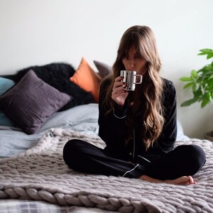 May include: A person in black pajamas with white trim sits on a bed, holding a silver mug. The bed is covered with a gray chunky knit blanket and pillows in various colors. A potted plant is in the background.