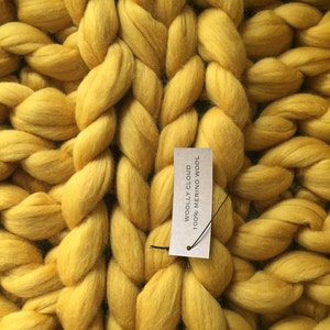 May include: Close-up of a chunky knit blanket in a vibrant yellow color. The blanket is made of thick, braided strands of wool. A tag reads "WOOLLY CLOUD 100% MERINO WOOL". The texture is soft and inviting, perfect for home decor.