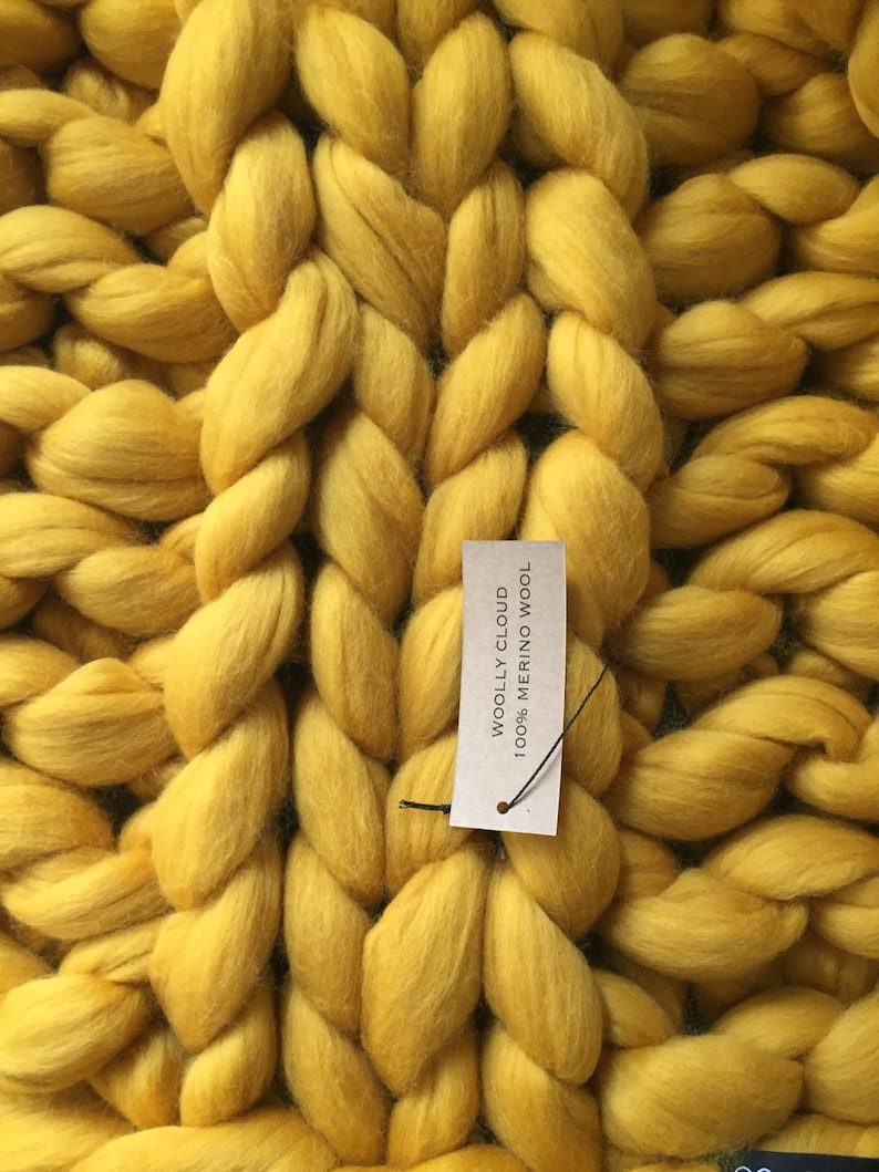 May include: Close-up of a chunky knit blanket in a vibrant yellow color. The blanket is made of thick, braided strands of wool. A tag reads "WOOLLY CLOUD 100% MERINO WOOL". The texture is soft and inviting, perfect for home decor.