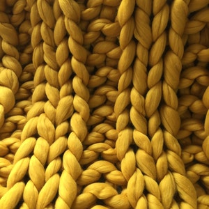 May include: Close-up of a chunky knit blanket in a mustard yellow color. The blanket is made of thick, braided yarn, creating a textured surface. The large stitches and warm color suggest a cozy, handmade item, perfect for home decor.