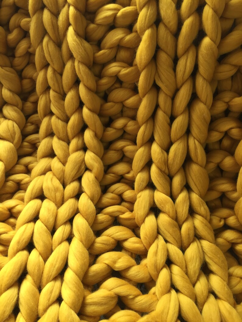 May include: Close-up of a chunky knit blanket in a mustard yellow color. The blanket is made of thick, braided yarn, creating a textured surface. The large stitches and warm color suggest a cozy, handmade item, perfect for home decor.