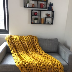 May include: A mustard yellow chunky knit blanket draped over a gray sofa. The blanket is made of thick, oversized yarn. Two black wall shelves hold decorative items like a red mug, a small owl figurine, and a potted plant.