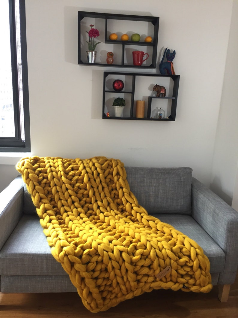 May include: A mustard yellow chunky knit blanket draped over a gray sofa. The blanket is made of thick, oversized yarn. Two black wall shelves hold decorative items like a red mug, a small owl figurine, and a potted plant.